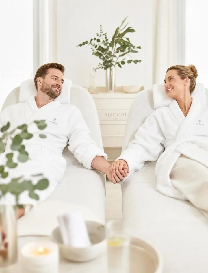 Couple in white spa robes holding hands in lounge chairs before a couples massage